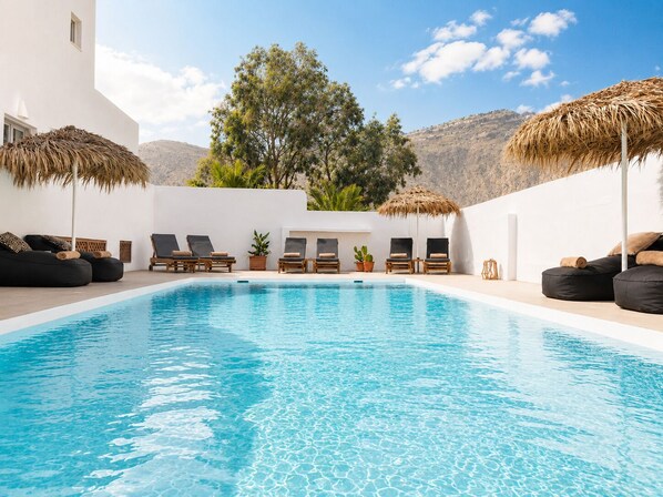 Un espace piscine doté de chaises longues et de parasols en chaume, entouré de murs blancs et bordé par un paysage montagneux.