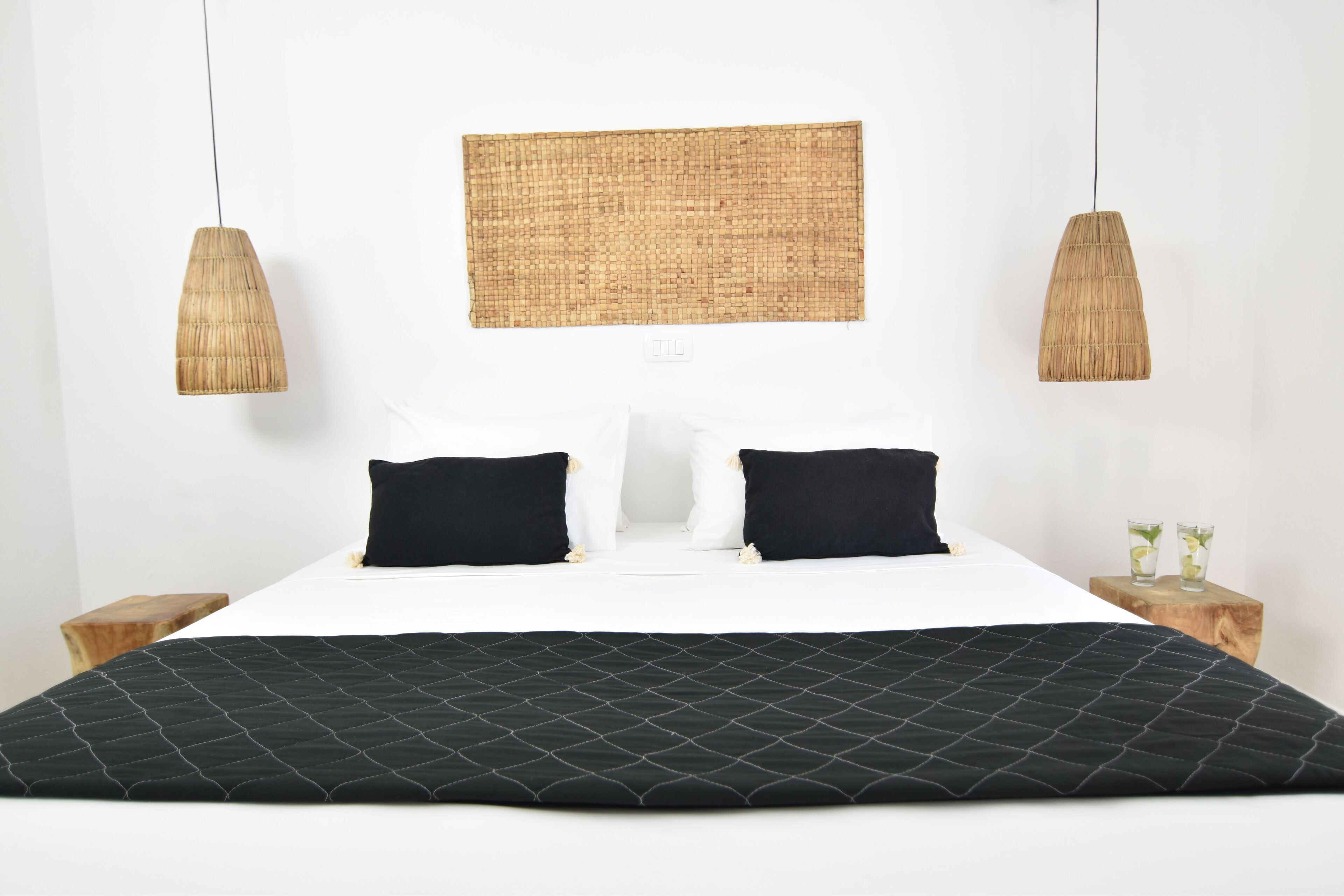 Una cama con una colcha acolchada negra, almohadas blancas y dos almohadas negras. Encima de la cama hay un tapiz de pared tejido y dos lámparas colgantes a cada lado.