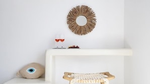 A minimalist room with a white bench, a woven stool, a decorative mirror, and a small table with a wine glass and grapes. - Aegeeis (Santorini)