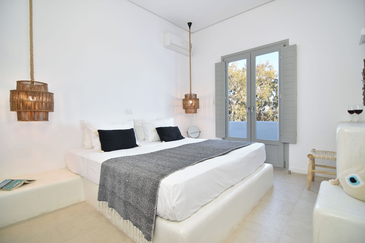 A bedroom with a large bed, two bedside tables, a window with shutters, and a hanging light fixture.