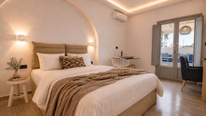 A modern bedroom with a large bed, bedside tables, a small table with a chair, and a view through a glass door. - Aegeeis (Santorini)