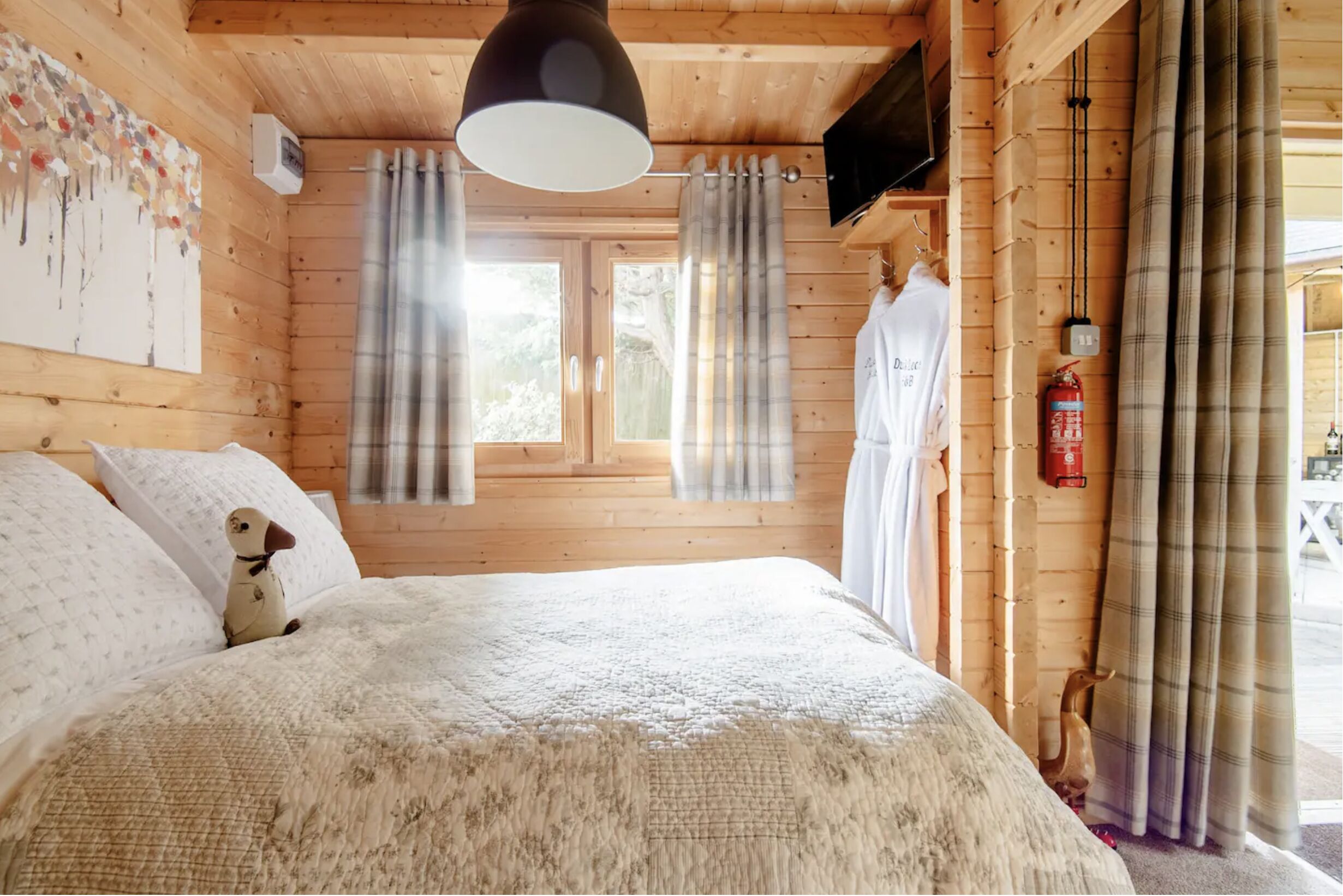 Een slaapkamer in een houten chalet, met een bed, een hanglamp, een brandblusser en een raam met gordijnen.