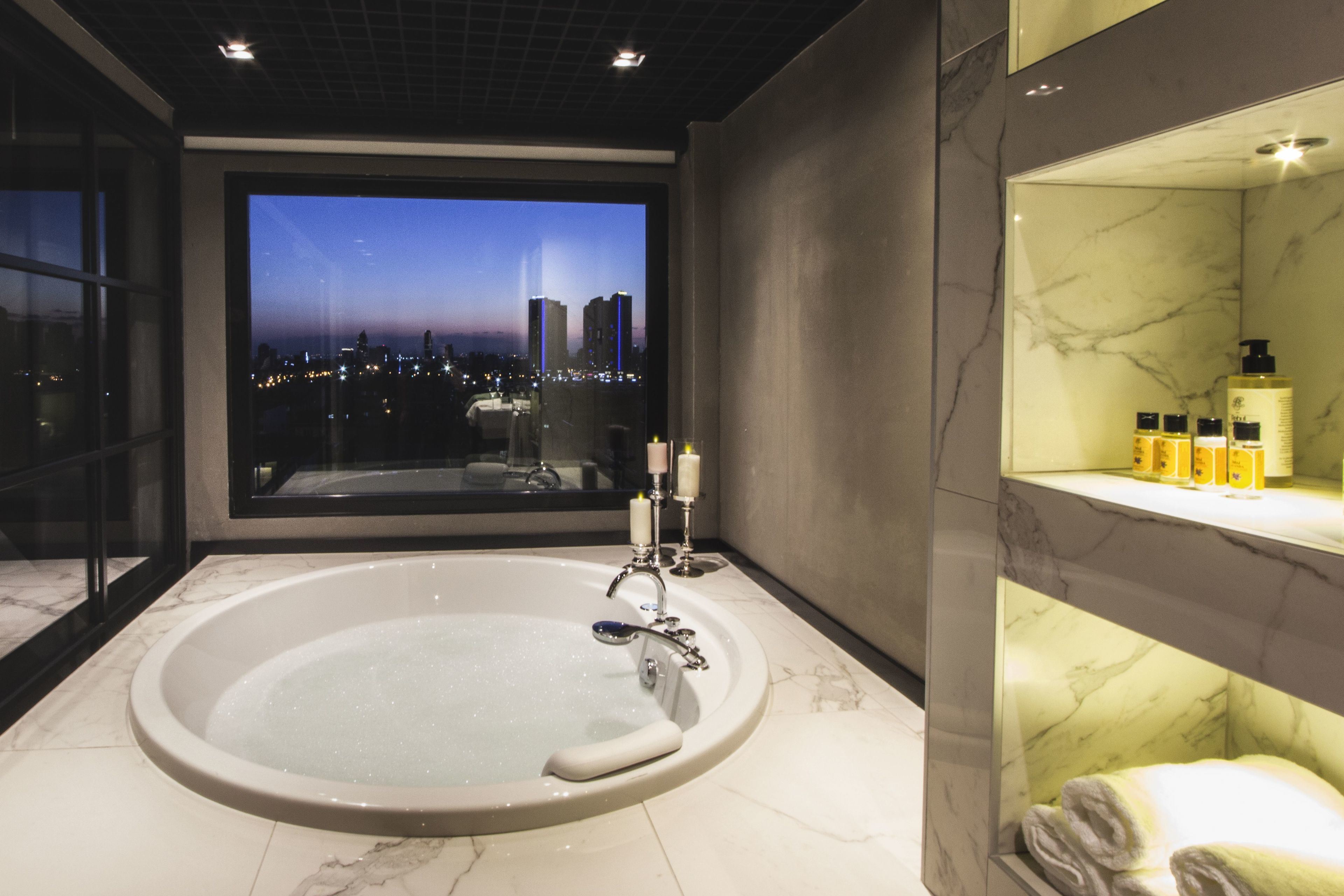 A modern bathroom with a large bathtub, a vanity with toiletries, and a view of a cityscape at dusk.