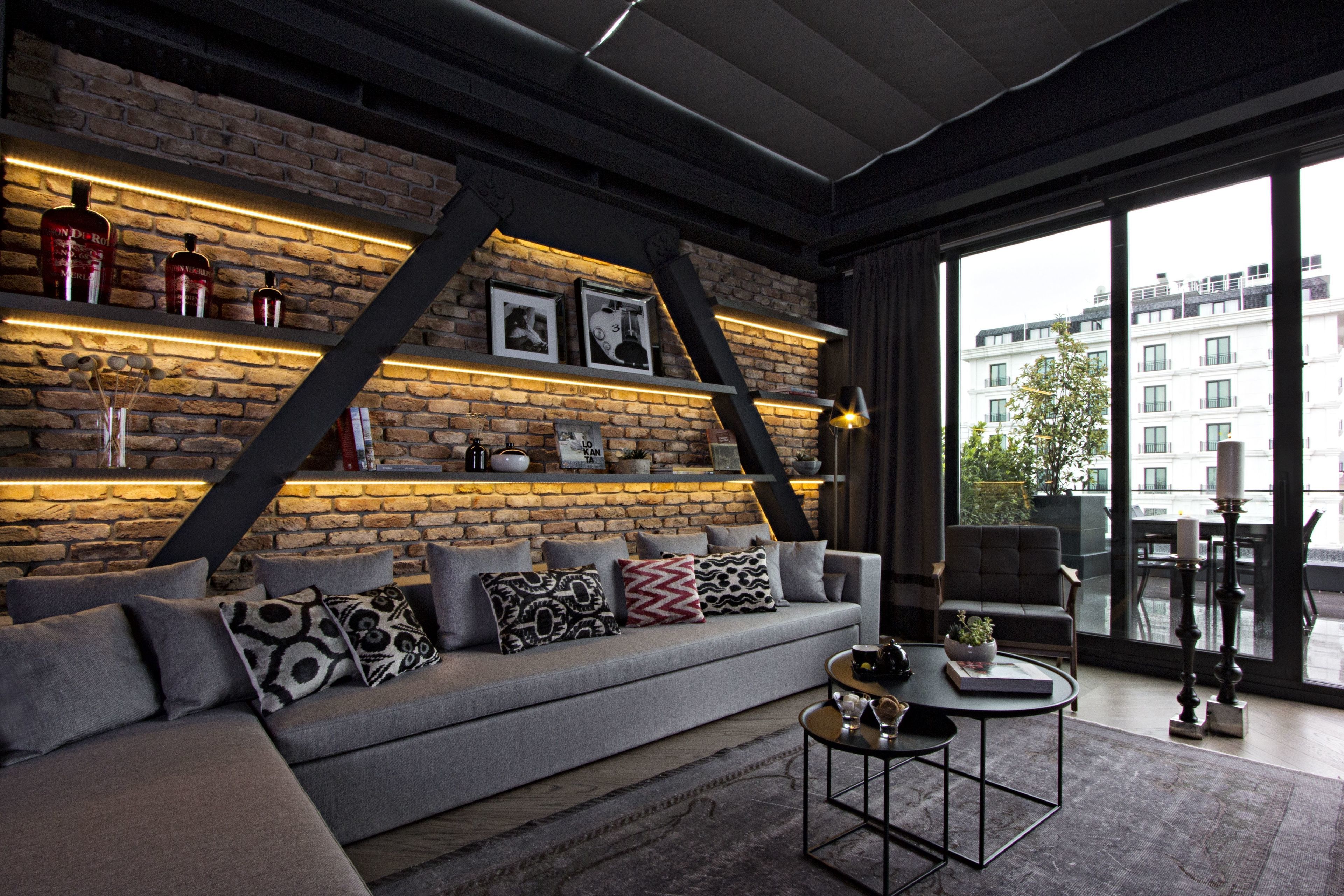 Una sala de estar moderna con una pared de ladrillo, un sofá gris con cojines estampados, una mesa de centro y vistas de edificios a través de amplios ventanales.