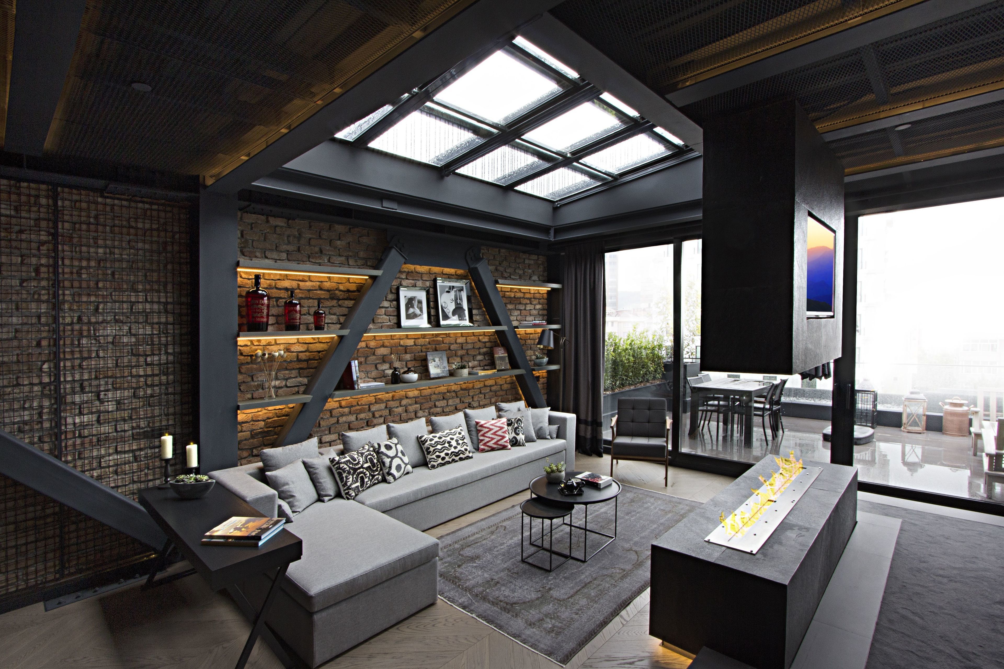 A modern living room with a grey sofa, a coffee table, and a fireplace. There is a brick wall and a large window with a view of the city.