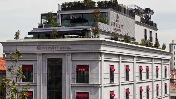A hotel with a classic facade, multiple windows with red awnings, and a rooftop terrace with greenery and satellite dishes.