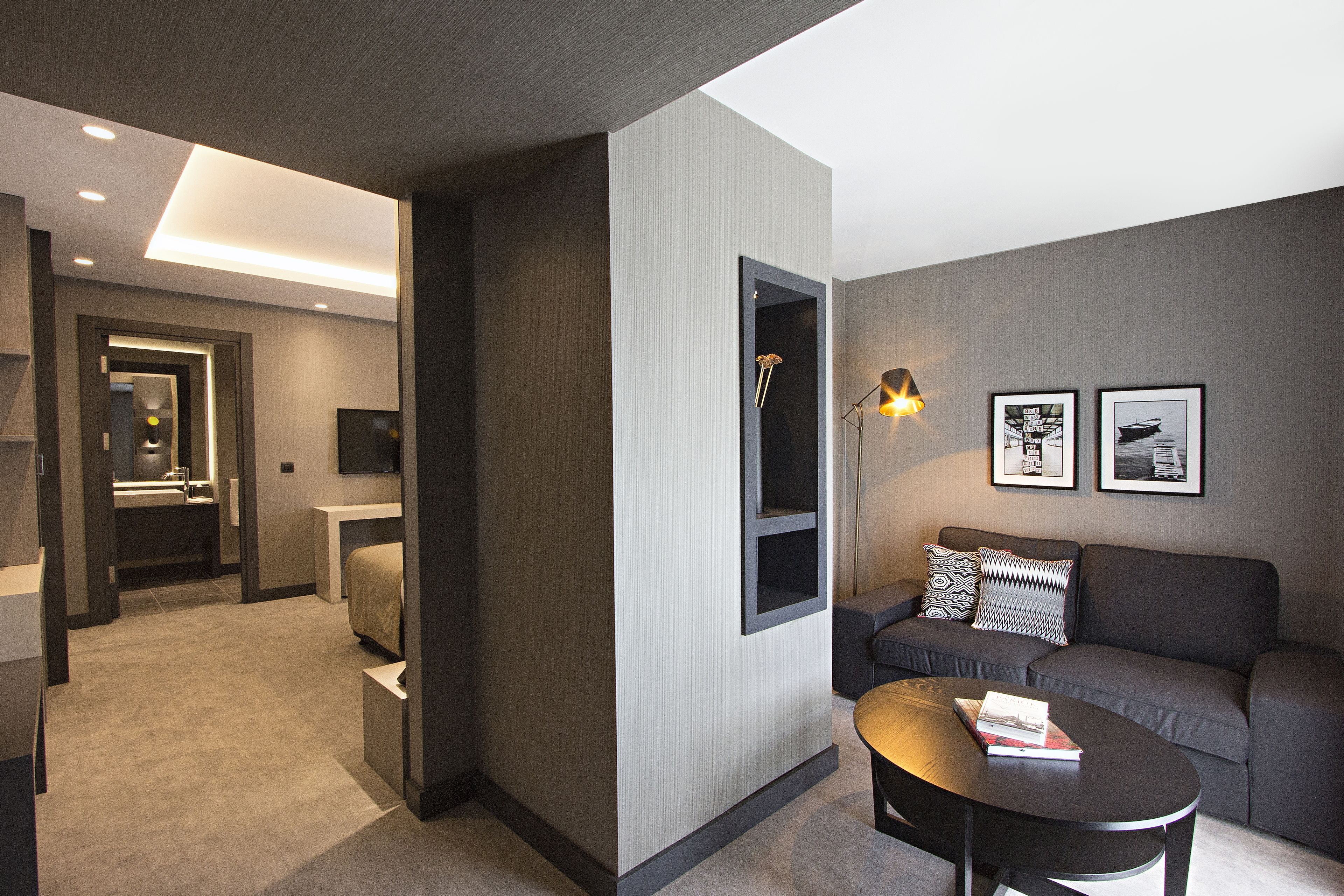 Et moderne hotelværelse med en sofa, et sofabord og et fjernsyn. Badeværelset har en vask og et spejl.