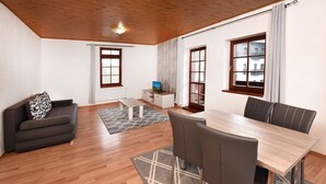 A modern living room with a sofa, a dining table with chairs, a TV on a cabinet, and large windows with curtains. - Villa Kunerad (Vysoké Tatry)
