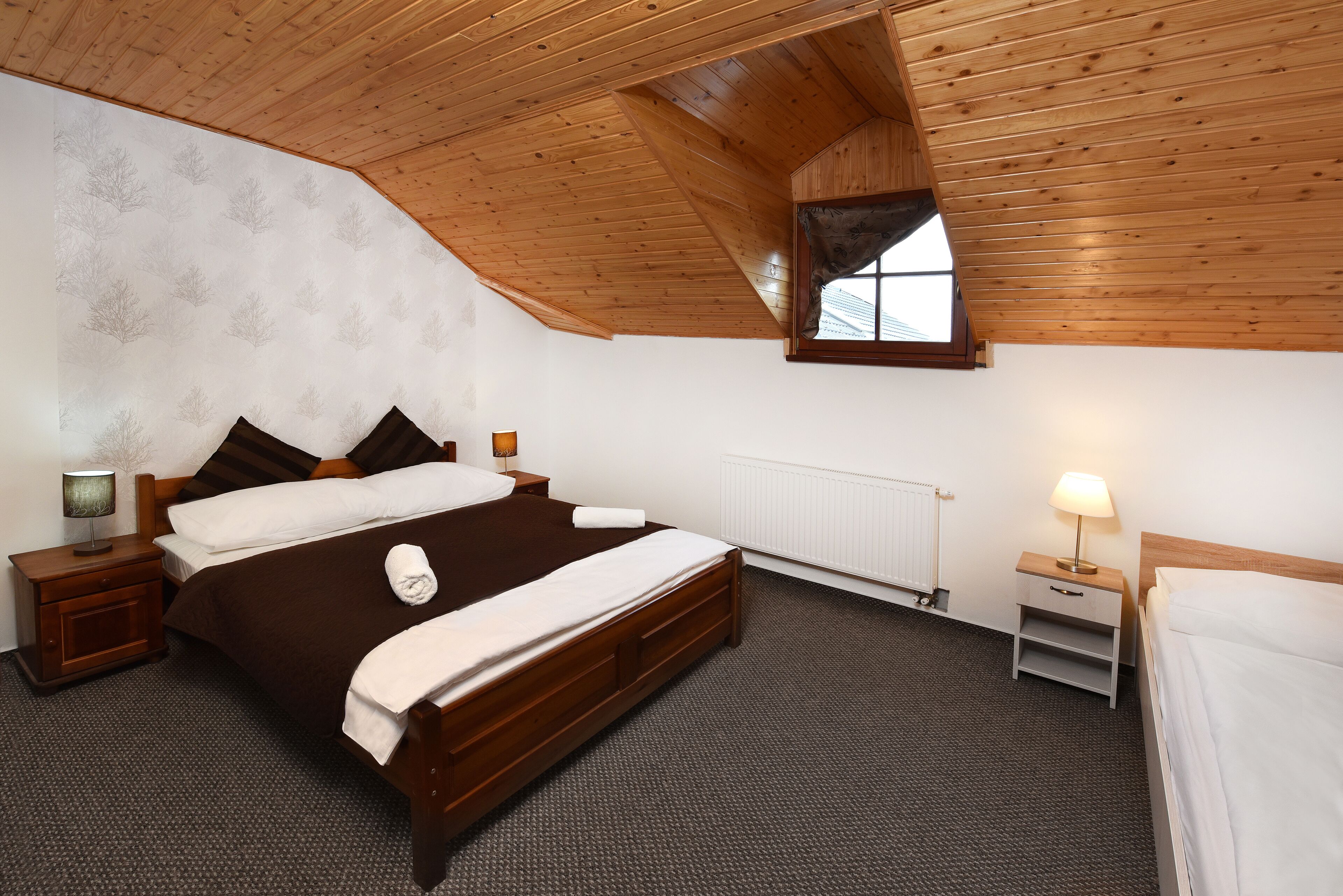 A bedroom with a wooden ceiling, a large bed with a brown bedspread, two nightstands with lamps, a window, and a radiator.