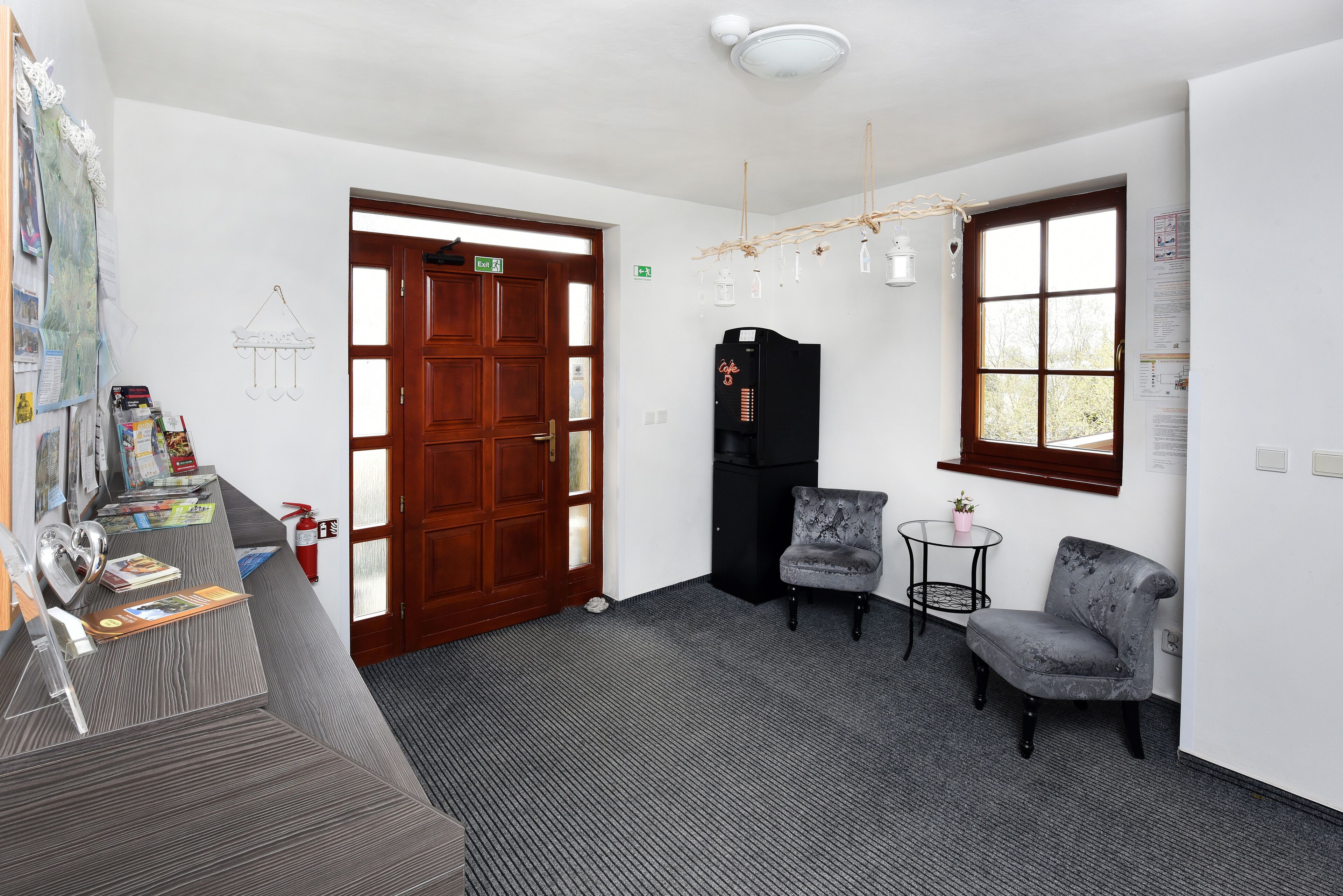 A reception area with a wooden door, a black refrigerator, two grey armchairs, a small table with a plant, and a wall-mounted light fixture.