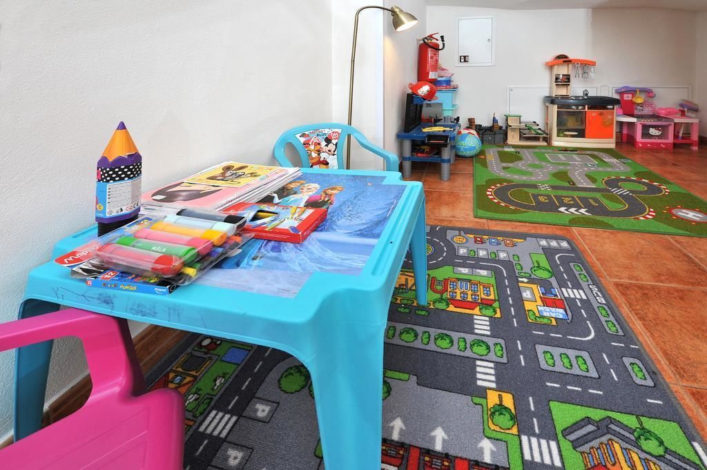 Una stanza giochi per bambini con un tavolo blu, sedie colorate, un tappetino da gioco e diversi giocattoli.