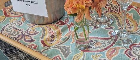 A champagne bottle in a metal bucket with a price tag, two empty wine glasses, and a vase with flowers on a patterned tablecloth.