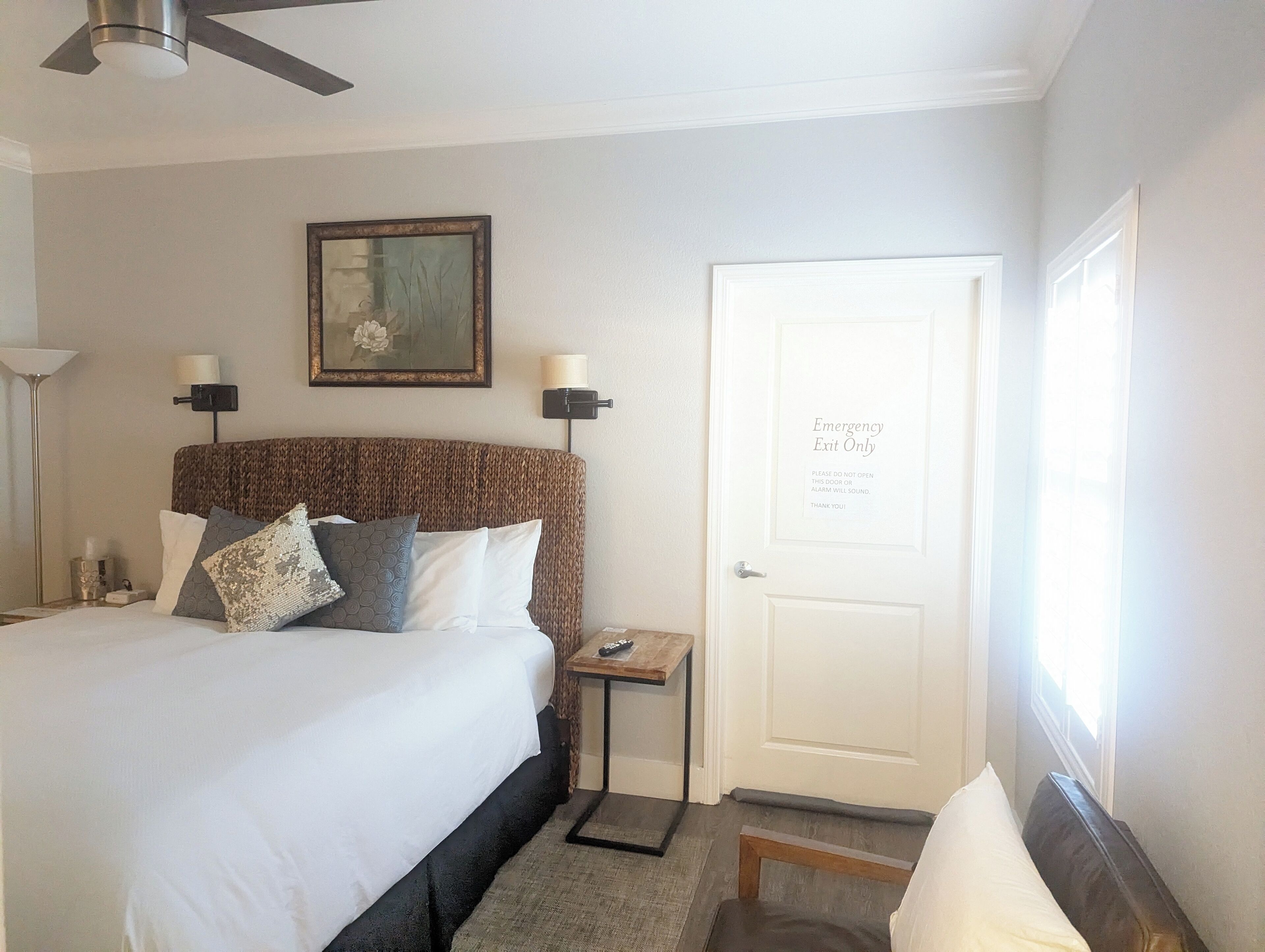 A hotel room with a large bed, a bedside table with a lamp, a framed picture on the wall, and a door with an emergency exit sign.