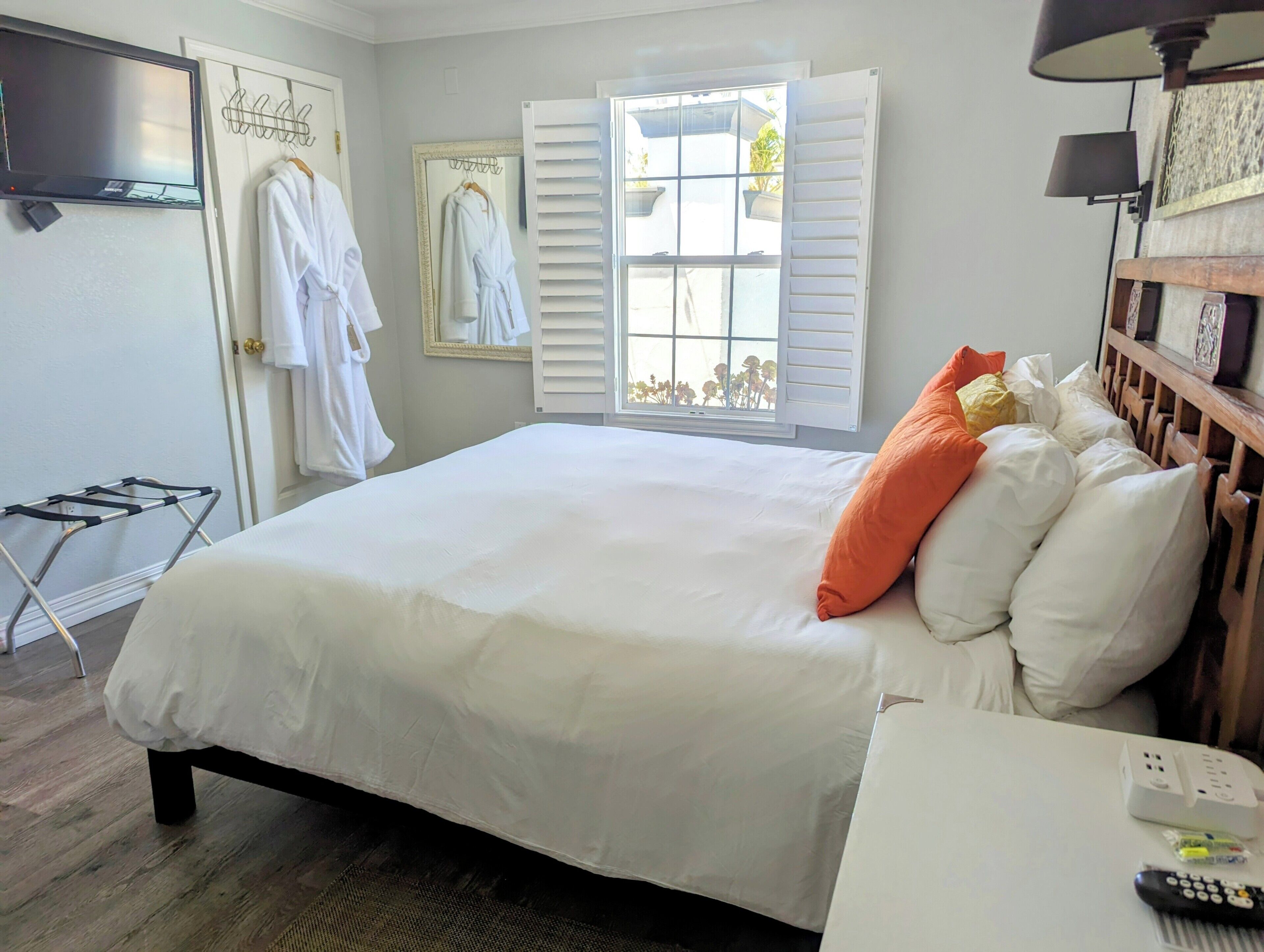 Un dormitorio con cama, ventana con contraventanas, televisor, tendedero y mesita de noche con control remoto.