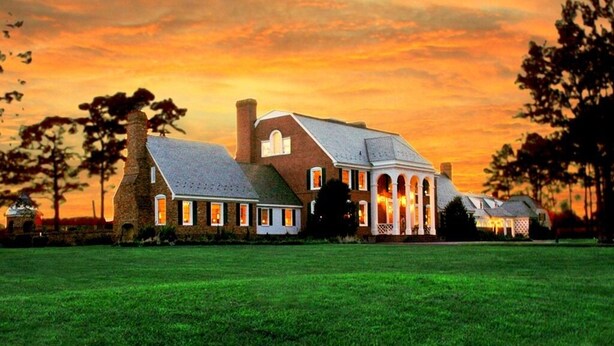 Een groot, historisch huis met meerdere schoorstenen, een ruim gazon en een zonsondergang.