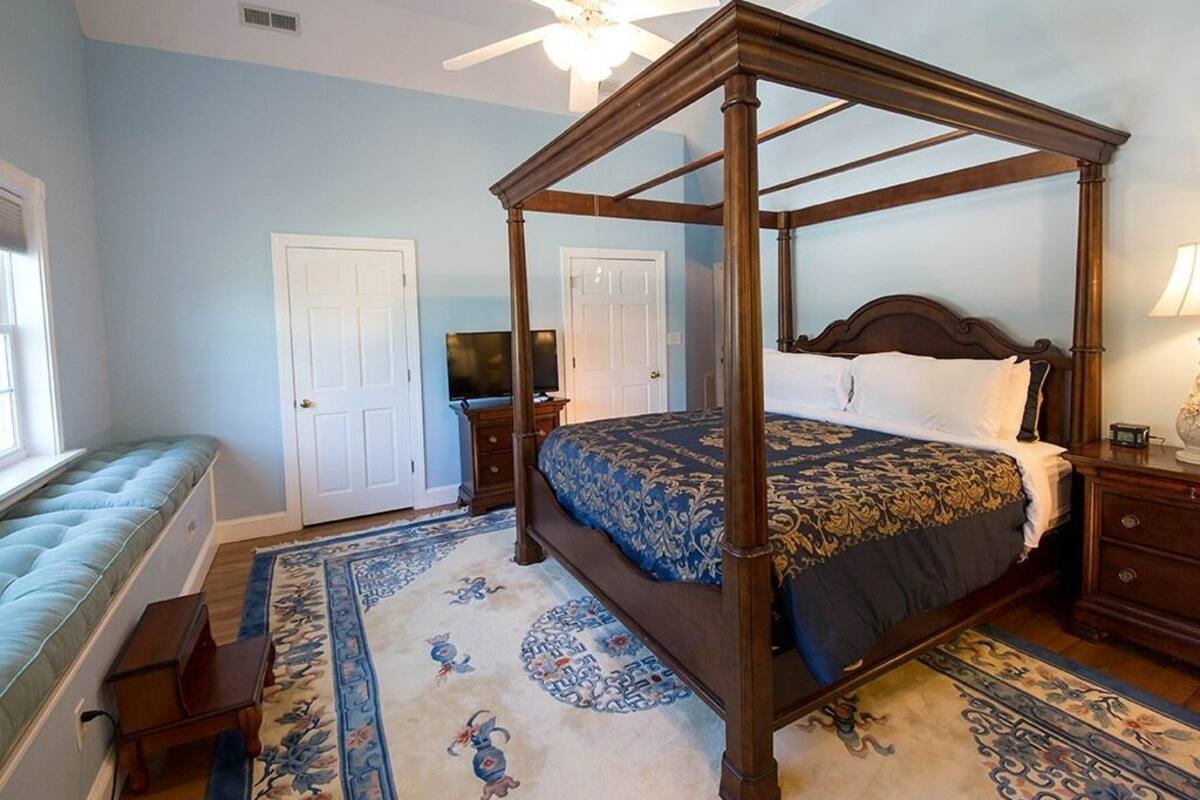 Ein Bett mit Baldachin, dessen dunkles Holzgestell einen blau-goldenen Federbettüberzug und ein passendes Kissen aufweist. Am rechten Rand des Bettes steht ein kleiner Nachttisch mit einer Lampe. Ein großer, kunstvoller Teppich mit floralen und tierischen Motiven bedeckt den Boden.