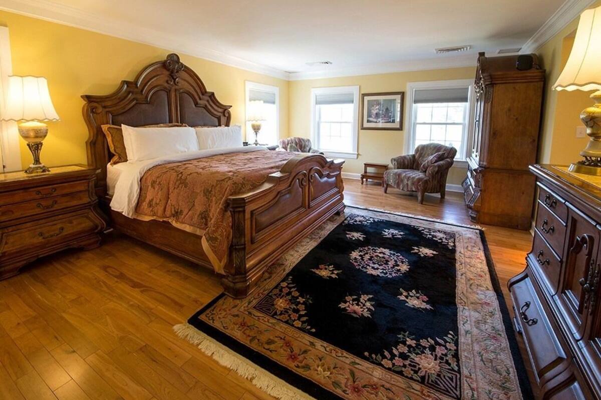 Ein Schlafzimmer mit einem großen Bett, einem Holz-Kopfteil, einem dunkel gemusterten Teppich, einer Holzkommode und einem Sessel.