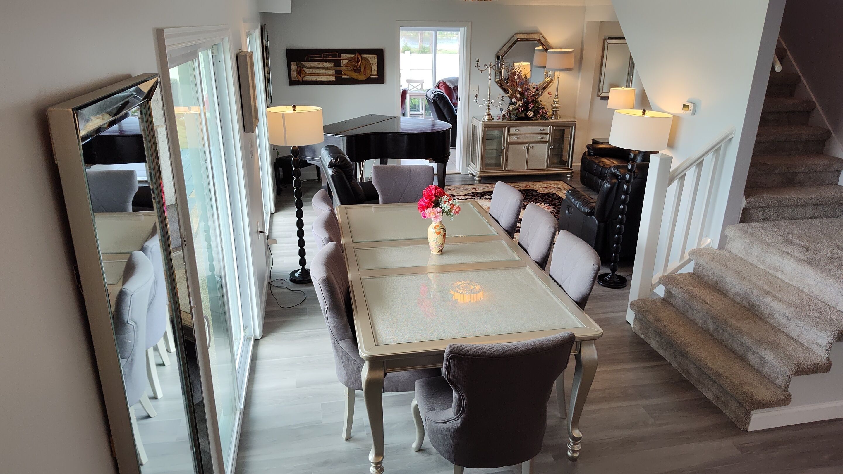 A dining area with a table, chairs, and a vase of flowers. There is a piano and a mirror in the background.