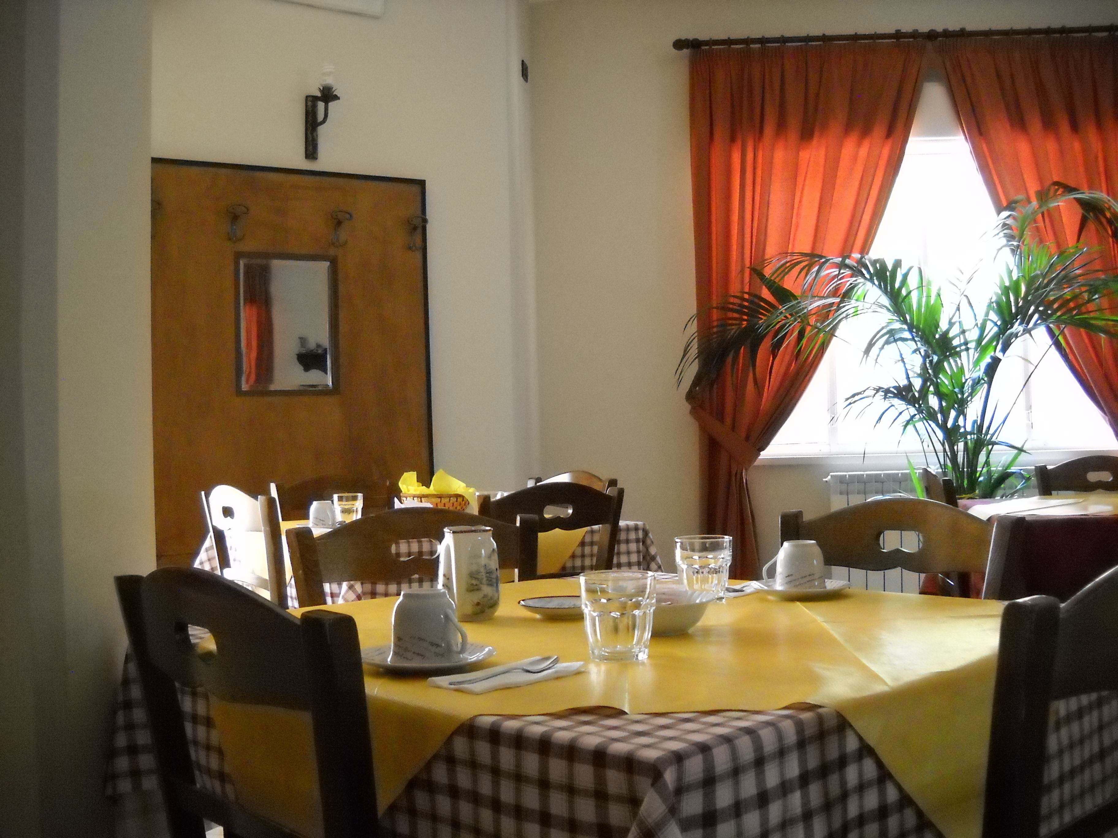 A dining area with a yellow table, checkered tablecloth, and chairs. There is a potted plant near the window, and a door with a mirror and a light fixture above it.