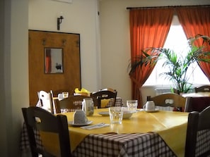 Un comedor con una mesa amarilla, mantel a cuadros y sillas. Hay una planta en maceta cerca de la ventana, y una puerta con un espejo y una lámpara arriba.