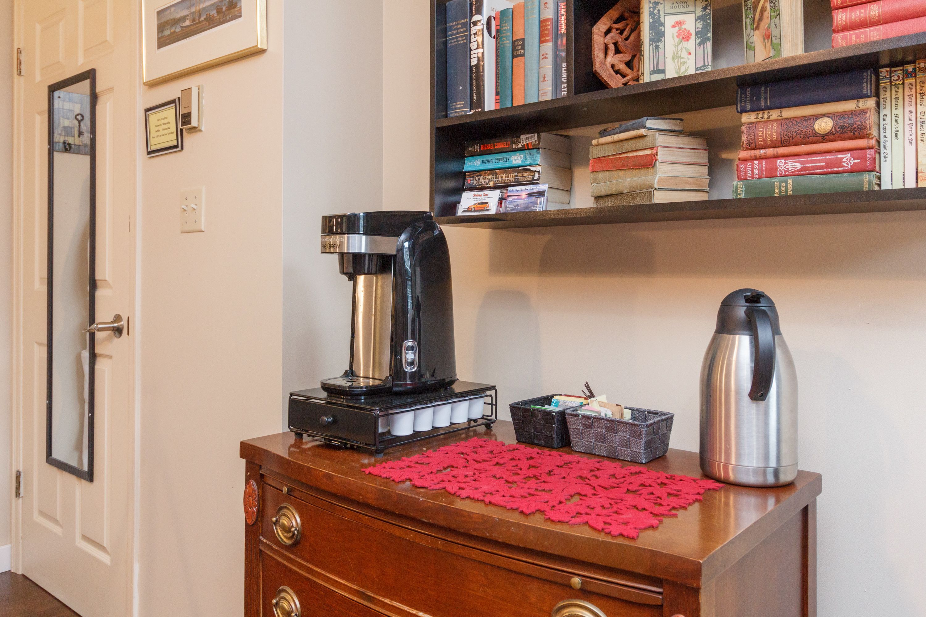 A coffee maker on a wooden dresser with a red doily, a bookshelf with books, and a door with a mirror.
