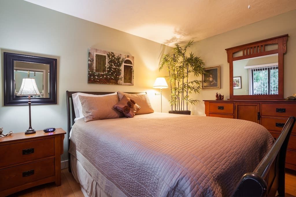 A bedroom with a large bed, a nightstand with a lamp, a dresser, and a mirror.