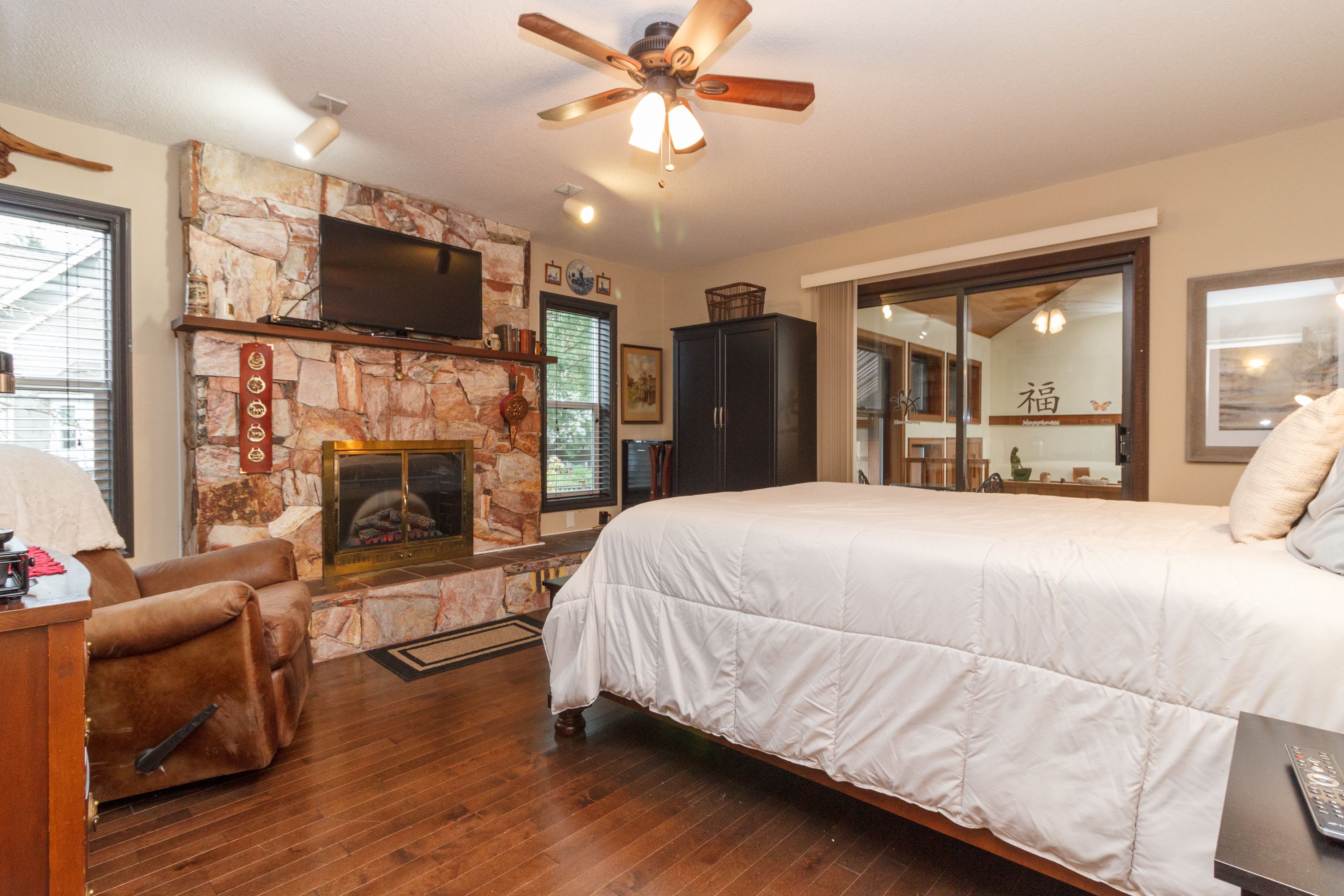 A bedroom with a large bed, a fireplace, a ceiling fan, and a television.