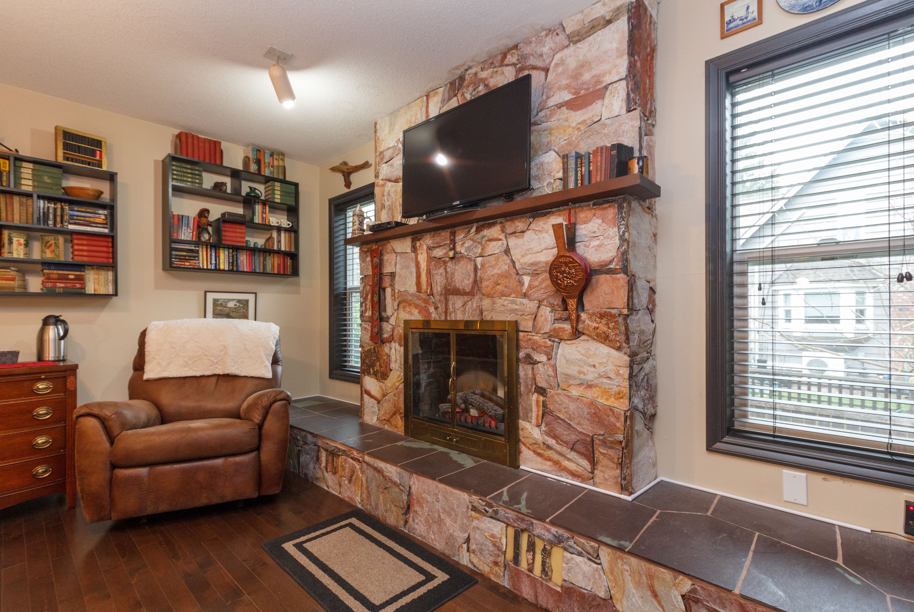 A living room with a stone fireplace, a leather armchair, a bookshelf, and a television mounted on the wall.