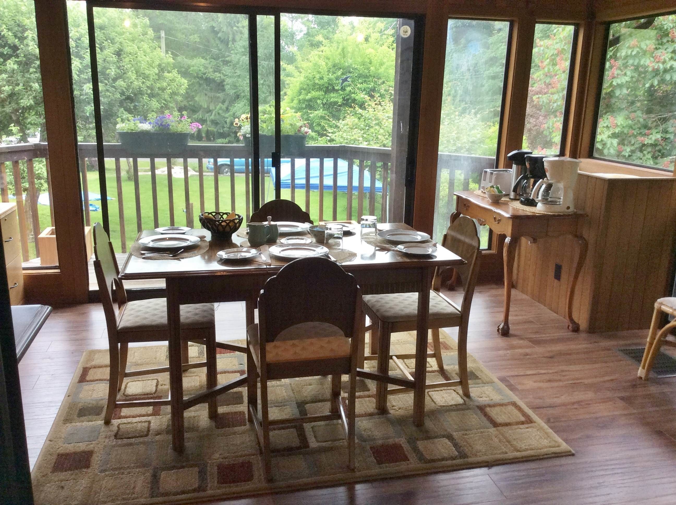 Un comedor con una mesa preparada para cuatro personas, un banco de madera y vista a un patio trasero con terraza de madera y follaje.