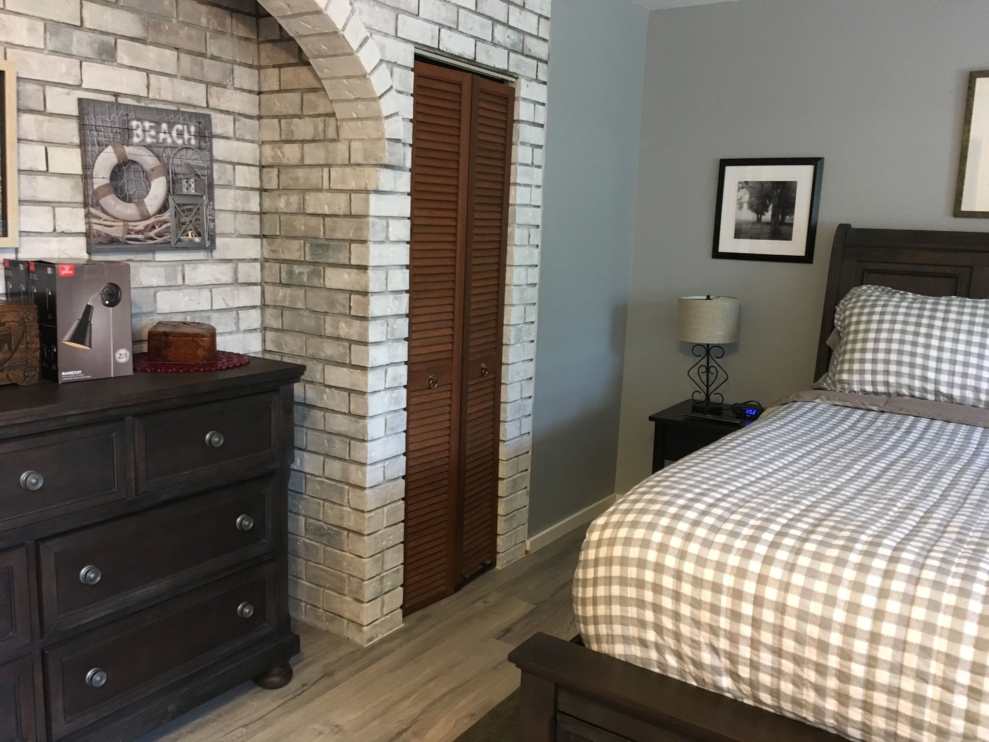 A bedroom with a brick fireplace, a wooden wardrobe, a bed with checkered bedding, a nightstand with a lamp, and a framed picture on the wall.