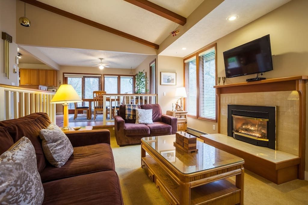 A cozy living room with a fireplace, a television, and a dining area in the background.