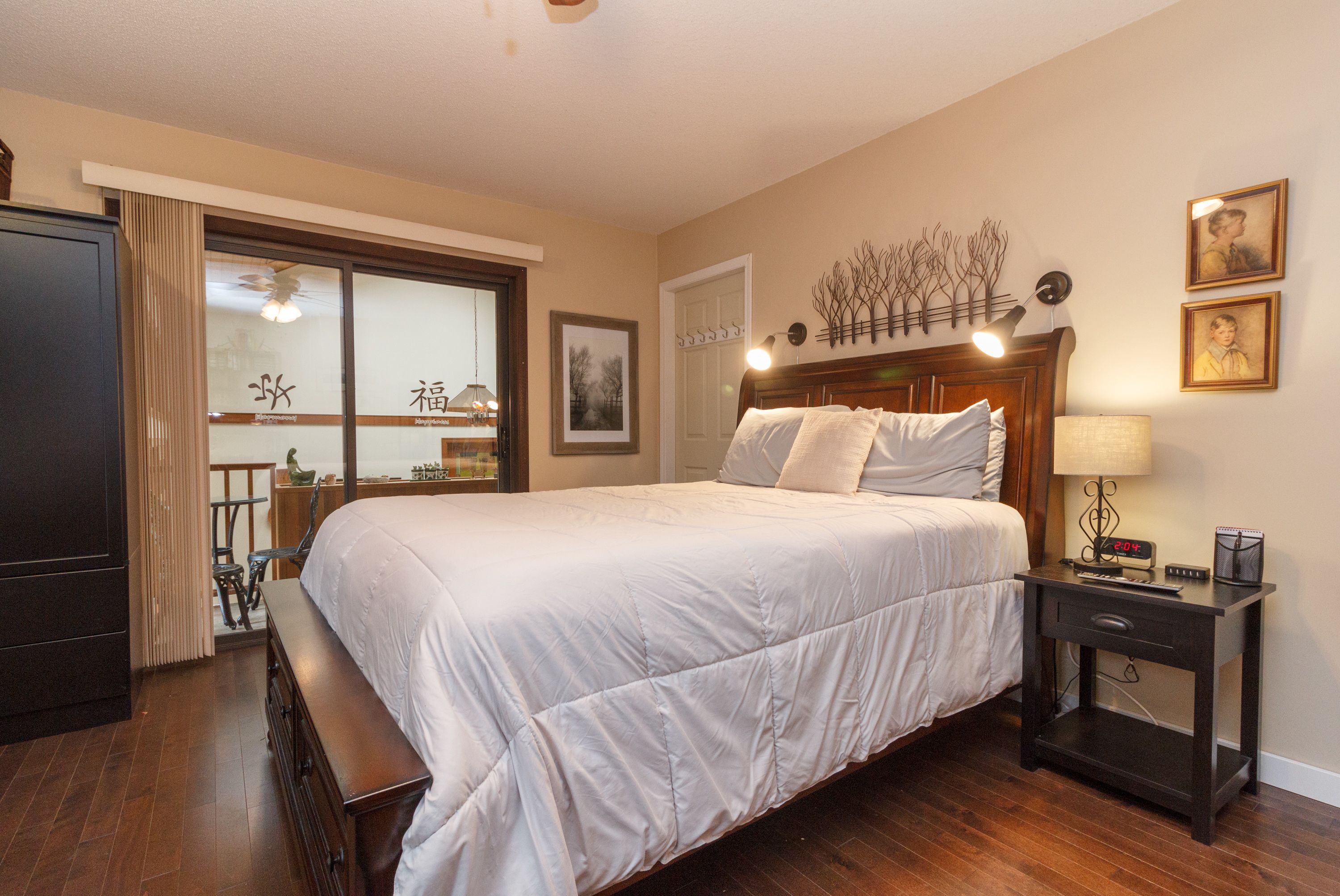 A bedroom with a large bed, a nightstand with a lamp, a wooden dresser, and a window with curtains.