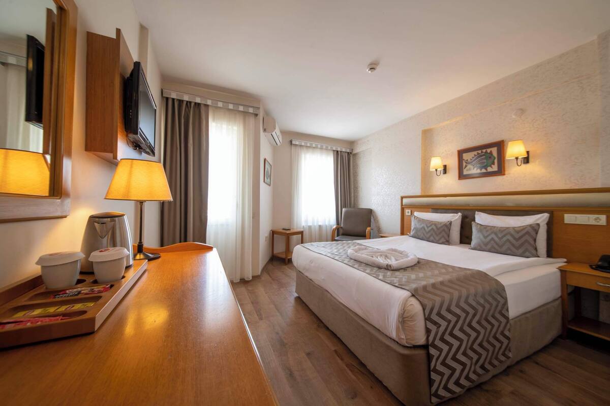A hotel room with a bed, bedside table, lamp, TV, and a small table with cups and a tray.