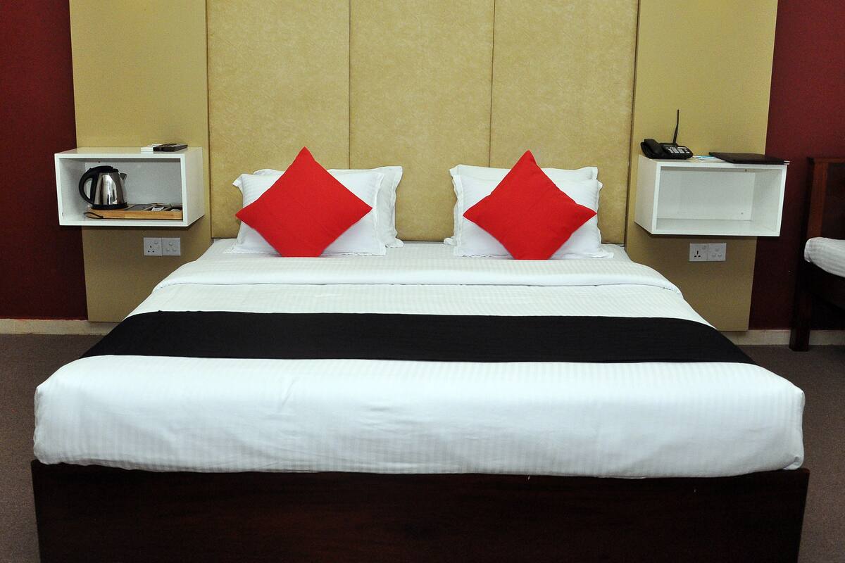 Une chambre d’hôtel avec un grand lit, deux oreillers rouges, une table de chevet blanche avec une bouilloire et un téléphone.