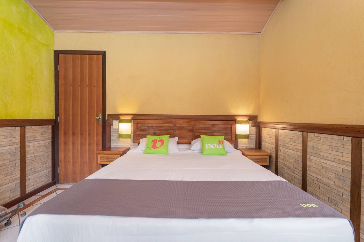 Un lit bien fait, avec une couverture marron et deux coussins verts ornés d’un « D », une tête de lit en bois et deux tables de chevet avec des lampes.