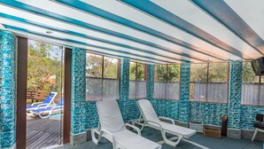 A sunroom with two white lounge chairs, a television, and a view of a pool area. - Wood House (Petrópolis)