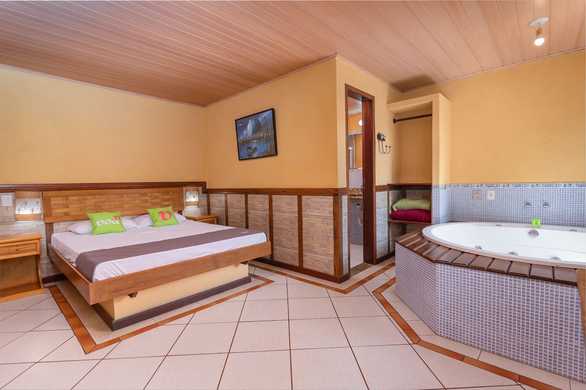 Une chambre à coucher avec un lit en bois, une petite table de chevet, une grande fenêtre et une salle de bain avec une baignoire.