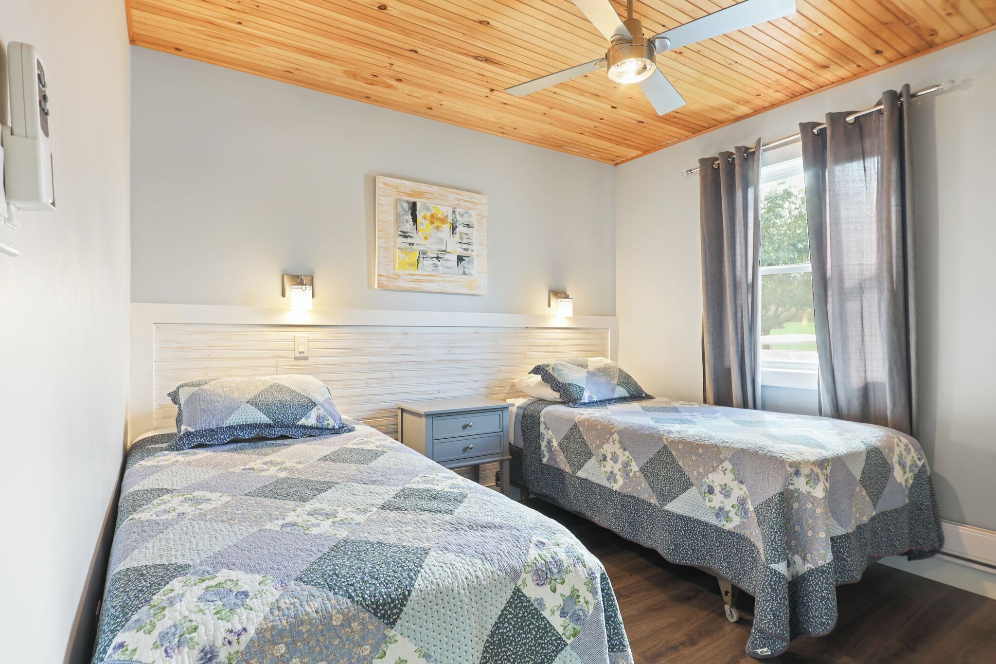 A bedroom with two beds, wooden ceiling, ceiling fan, window with curtains, and wall-mounted light fixtures.