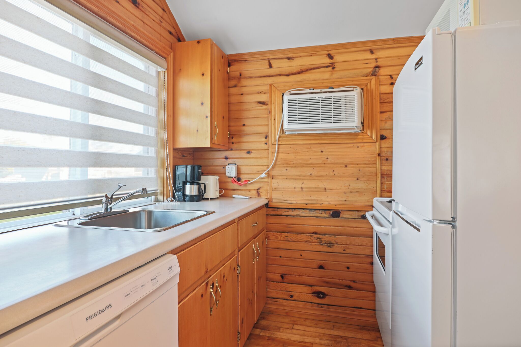 A compact kitchen with wooden walls, a white refrigerator, a built-in oven, a sink, and a window with blinds.