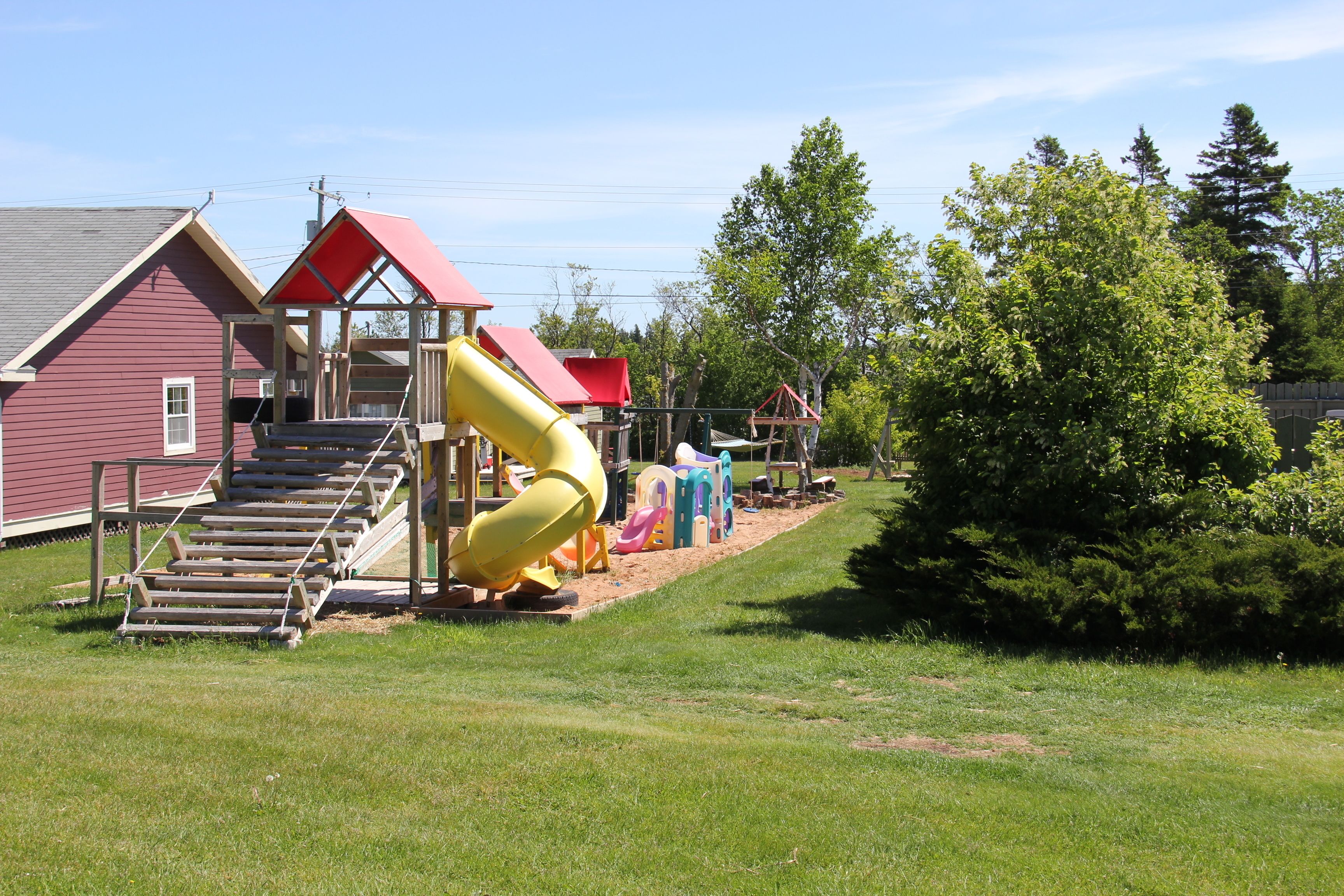 Une aire de jeux avec un toboggan jaune, une structure surmontée d’un toit rouge et un escalier en bois.