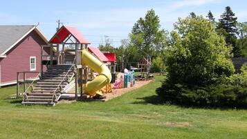 Une aire de jeux avec un toboggan jaune, une structure surmontée d’un toit rouge et un escalier en bois.