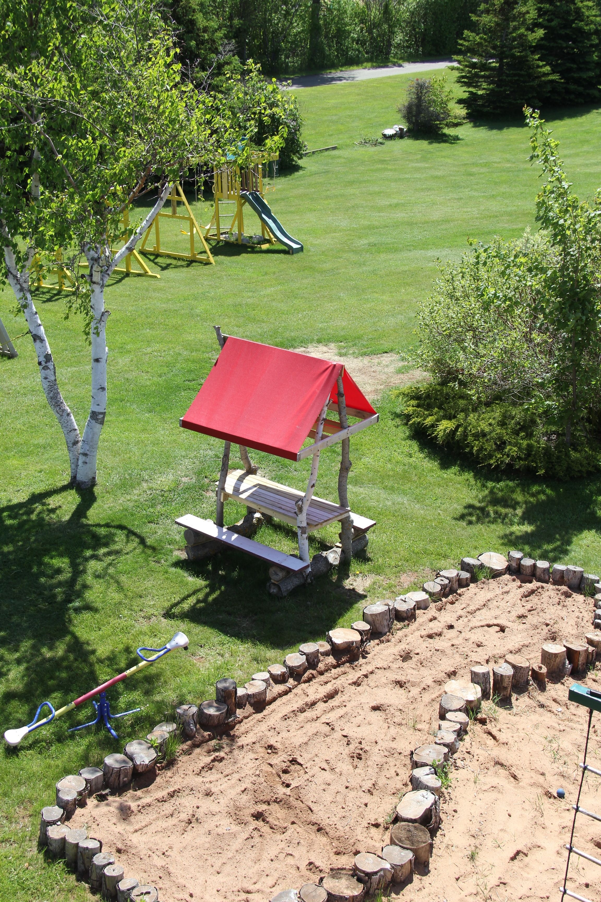 A playground with a slide and swings, surrounded by a grassy area with trees and a small wooden structure.