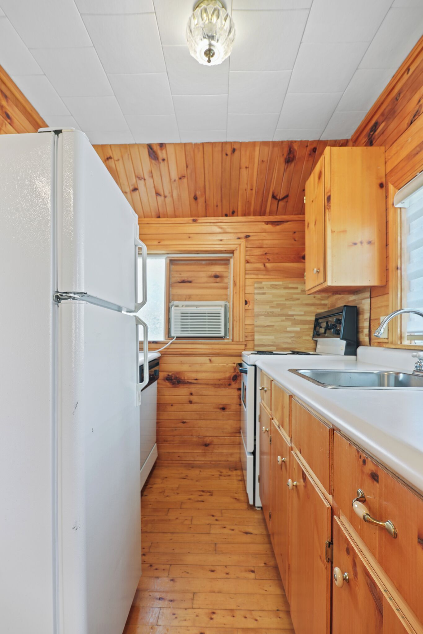 A compact kitchen with wooden walls, a white refrigerator, a microwave, a stove, and a sink.