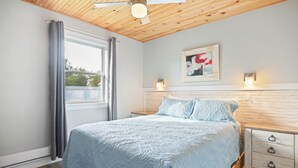 Un dormitorio con cama, ventilador de techo, ventana con cortinas, mesita de noche y luz de pared.