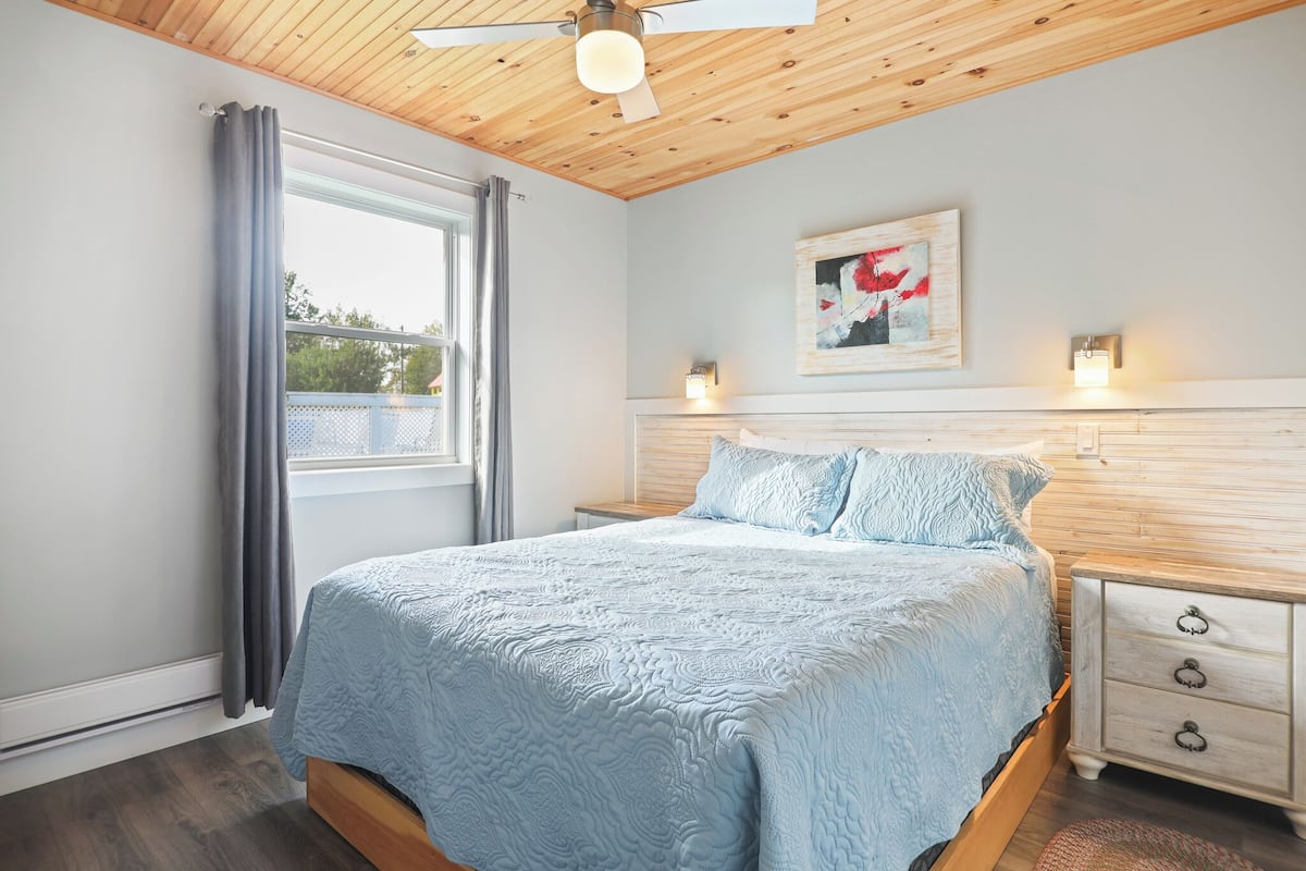 Ein Schlafzimmer mit einem Bett, einem Deckenventilator, einem Fenster mit Vorhängen, einem Nachttisch und einer Wandleuchte.
