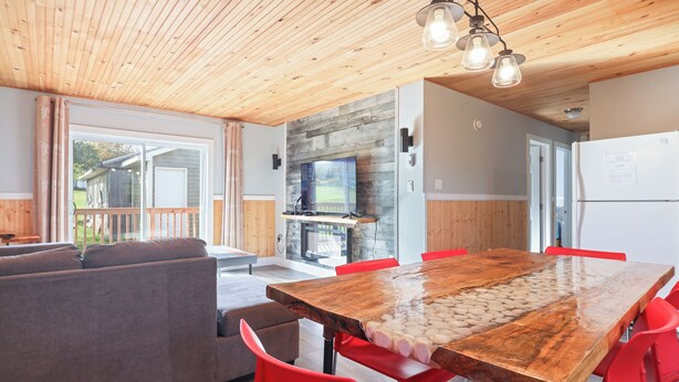 Una sala de estar acogedora con techo de madera, una amplia mesa de comedor de madera con sillas rojas, un sofá gris y una chimenea de piedra.