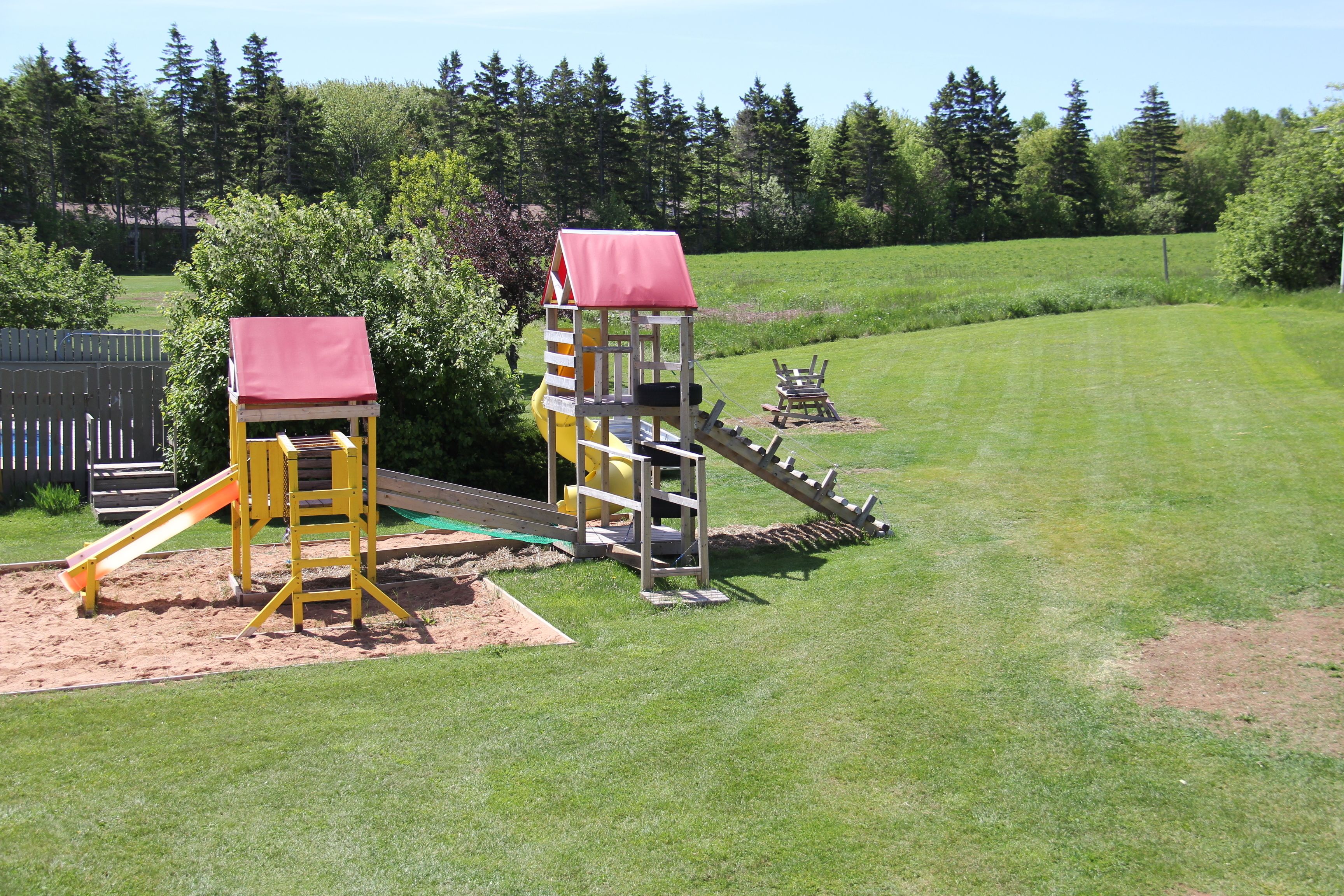 A playground with two play structures, including slides and climbing areas, surrounded by a grassy field and trees.