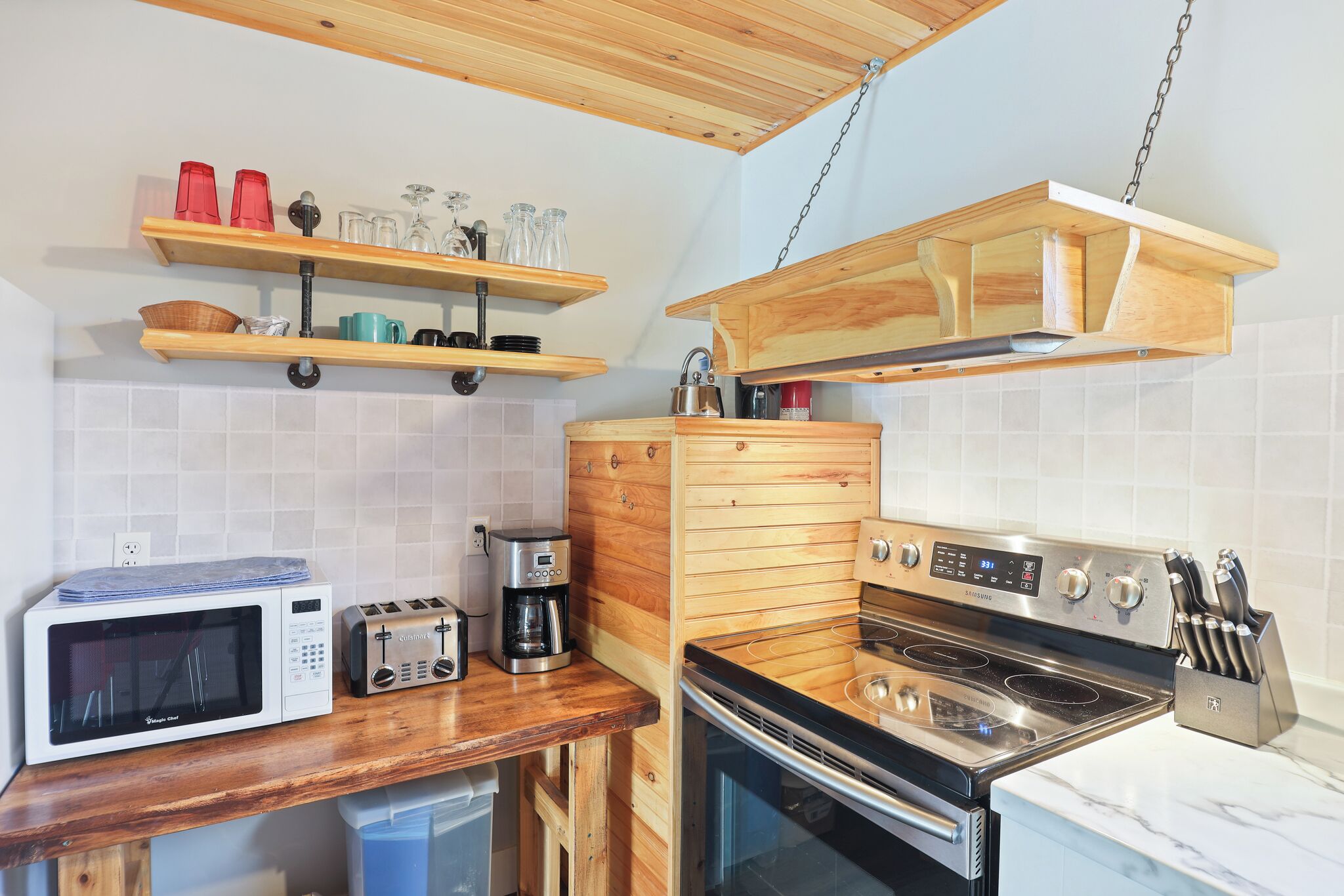A compact kitchen with a microwave, toaster, coffee maker, and stove. Wooden shelves and cabinets are present.