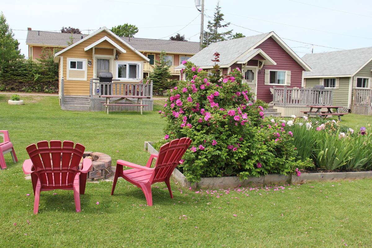 Eine Rasenfläche mit zwei roten Adirondack-Stühlen, einer Holzterrasse und einem Blumenbusch.
