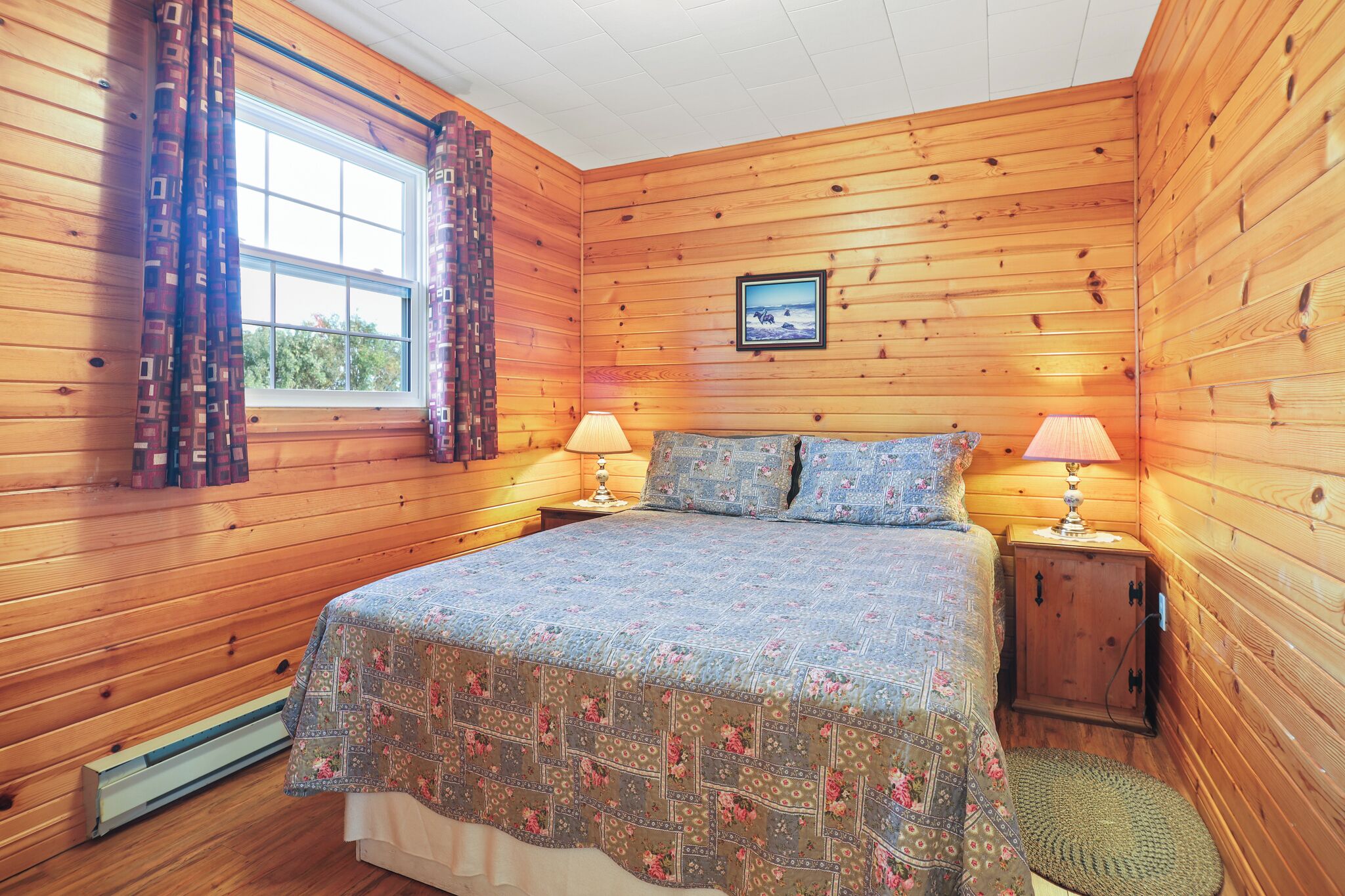 A wooden-paneled bedroom with a bed, two bedside lamps, a window with curtains, and a framed picture on the wall.